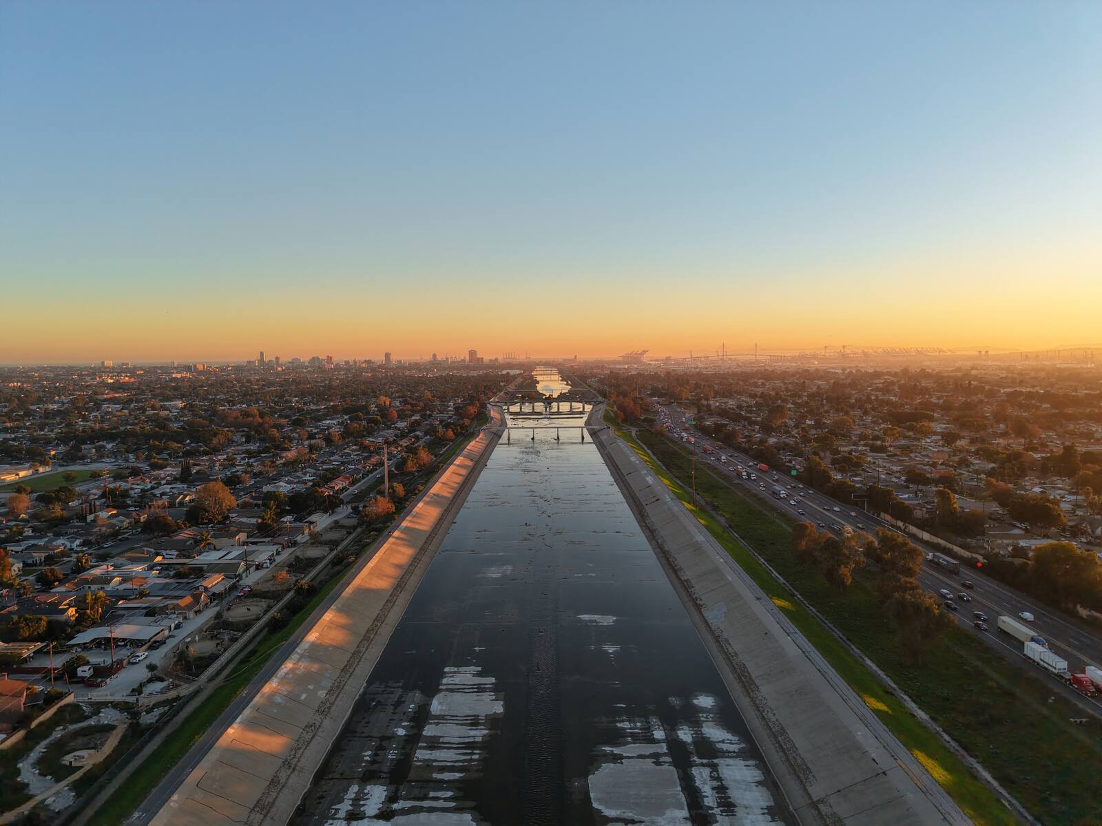 LA River channel at sunset — aerial view looking south along the concrete channel toward the Long Beach skyline at golden hour
