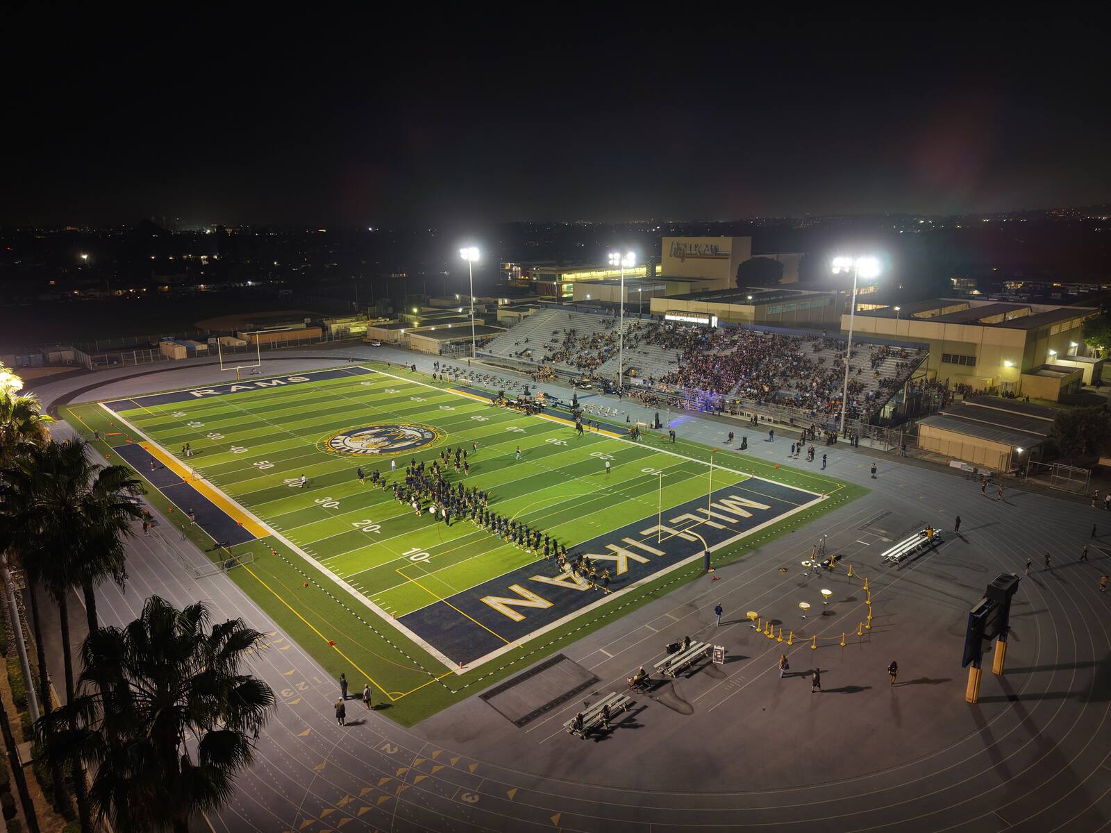 Millikan High School stadium at night — aerial view of the lit field with band on the turf and crowd in the stands