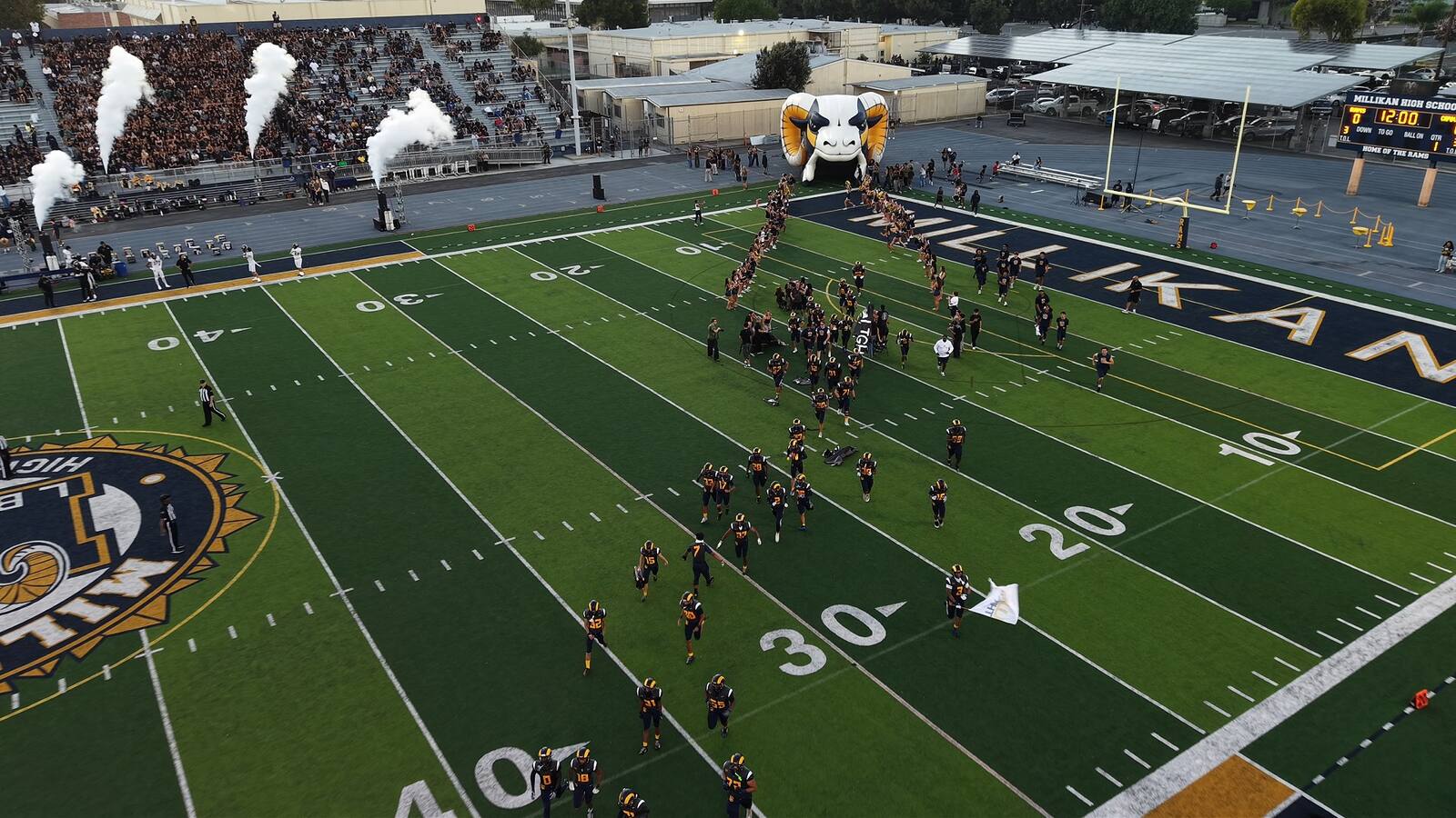 Millikan High School football game day — aerial view of the team running onto the field with smoke cannons firing and packed stands